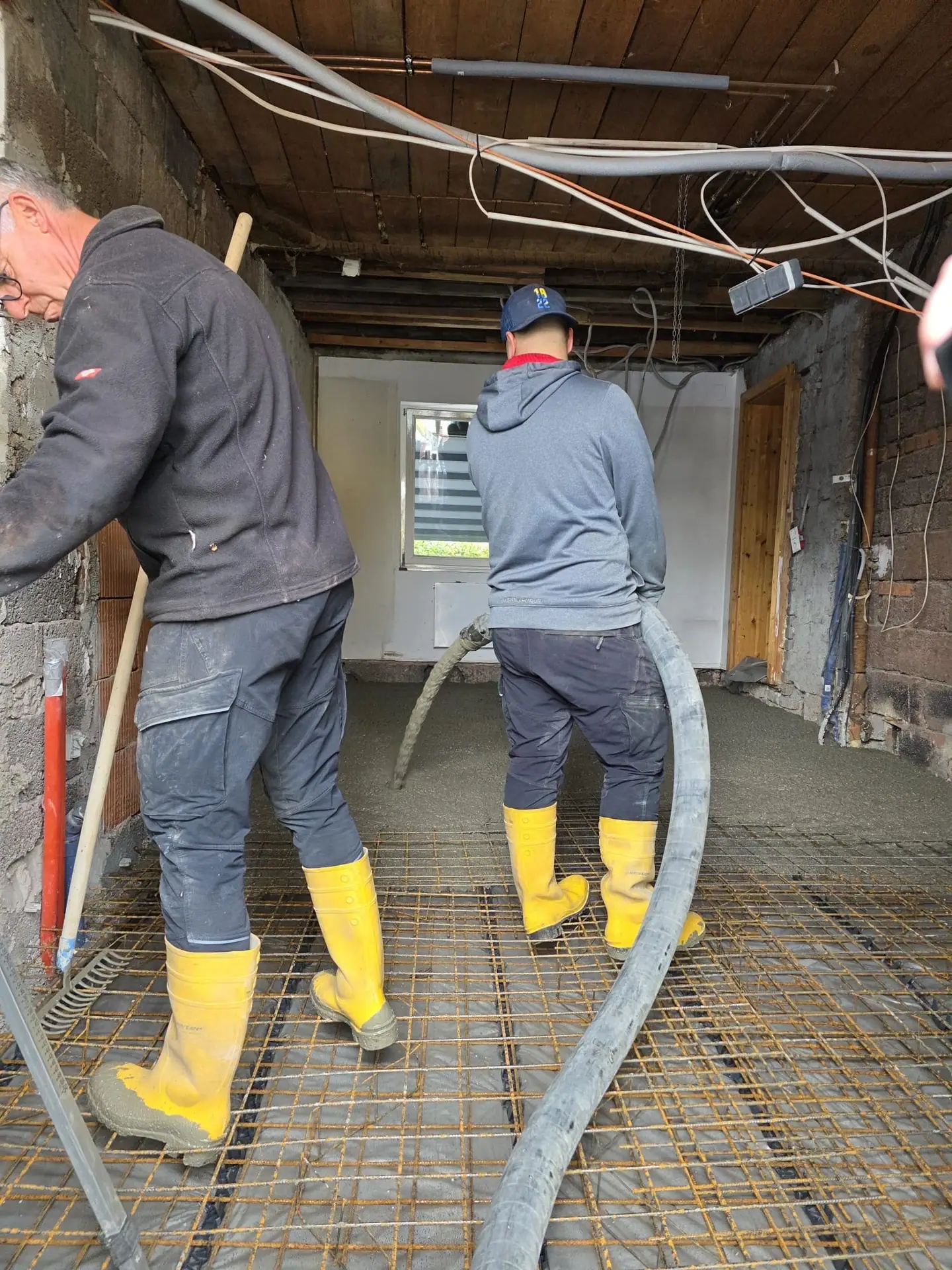 Zwei Arbeiter in Gummistiefeln bringen Estrich über einen Schlauch ein, unter einer Holzbalkendecke
