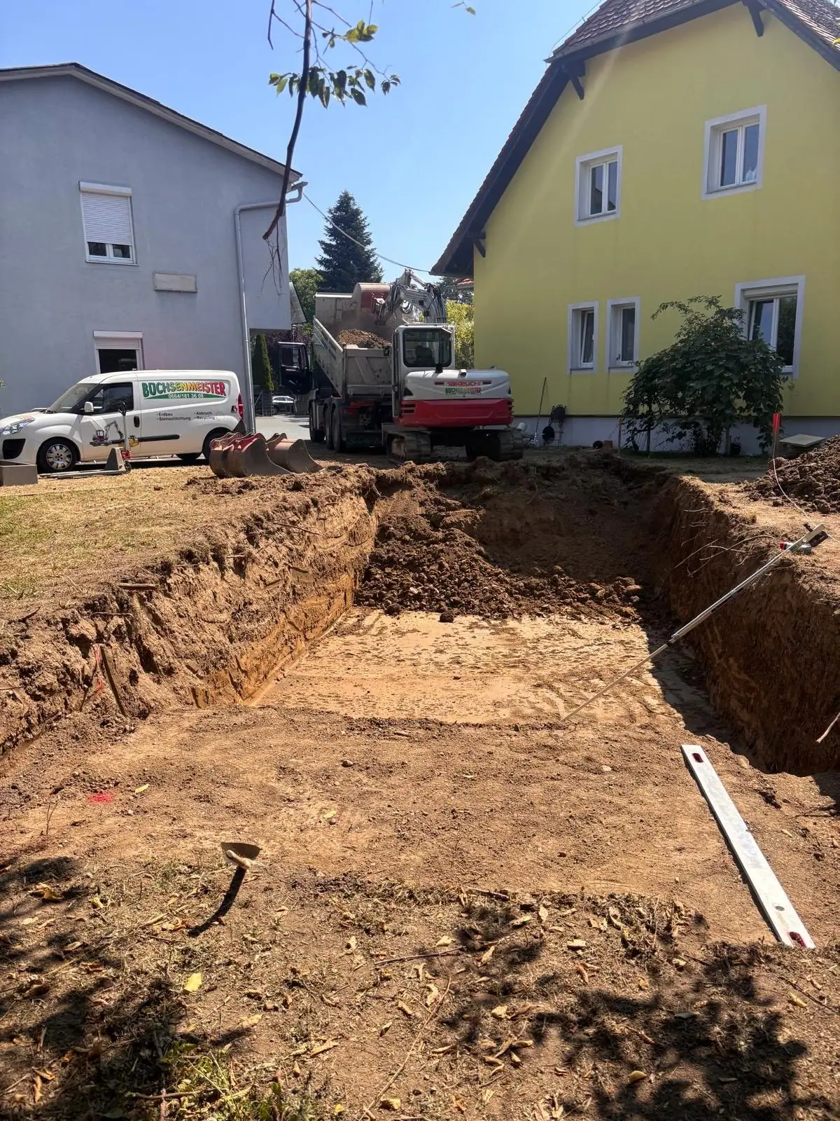 Ausgehobene Baugrube im Garten vor einem gelben Haus, dahinter Bagger und Lkw