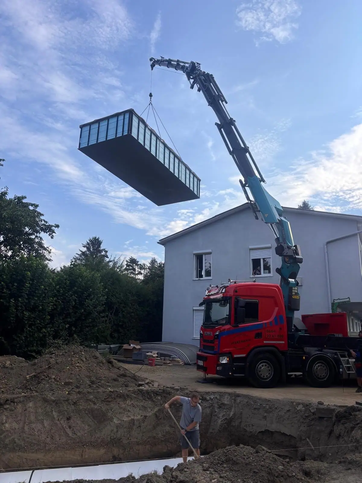 Kranwagen hebt ein Poolbecken durch die Luft, unten wartet ein Arbeiter in der Grube