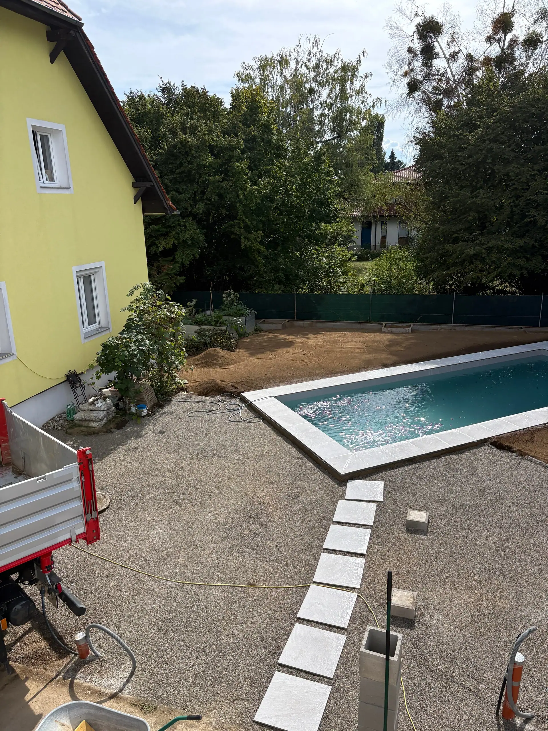 Pool mit heller Einfassung und Trittplatten neben einem Wohnhaus, dahinter Garten und Bäume