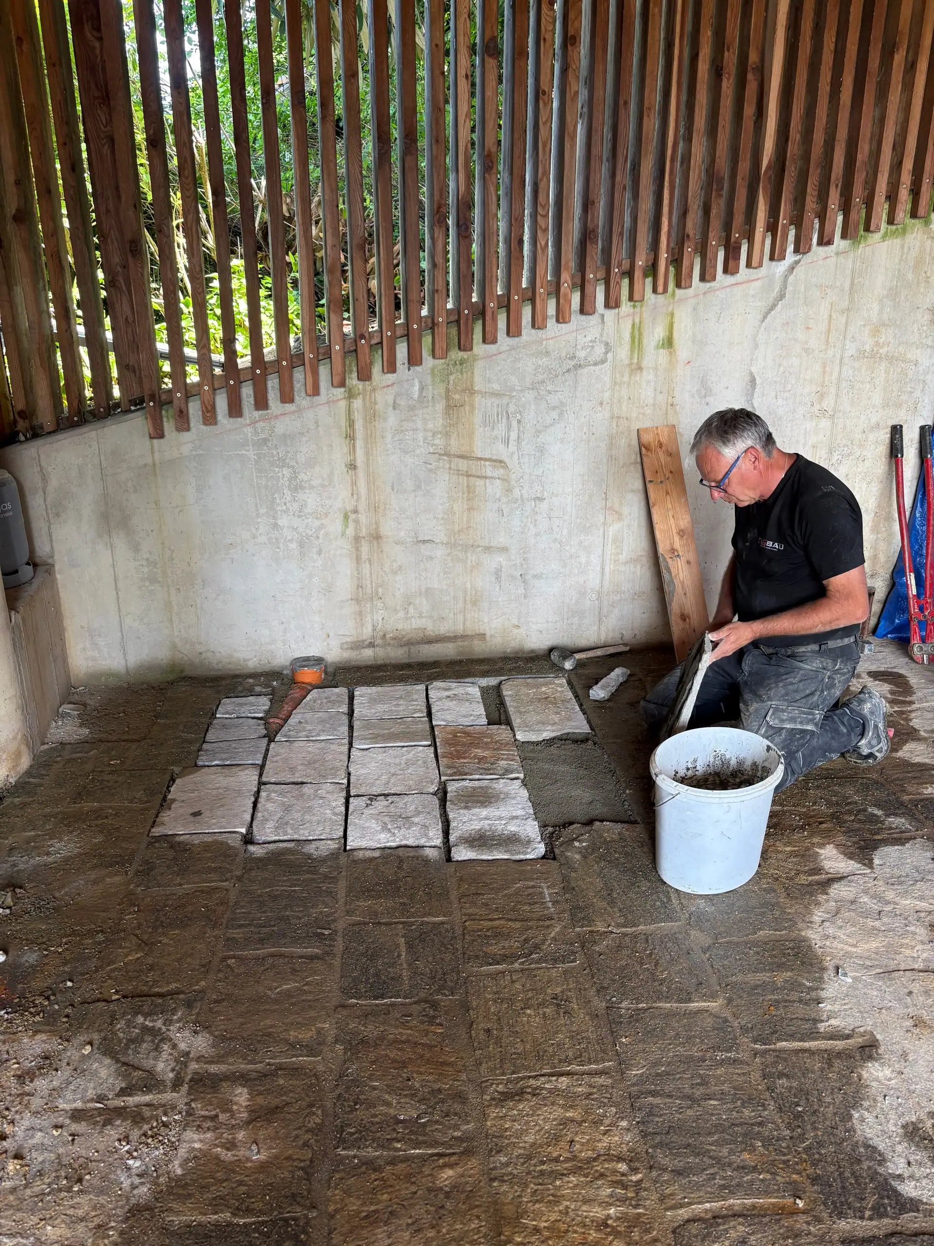 Arbeiter verlegt kniend Natursteinplatten, daneben ein Mörtelkübel